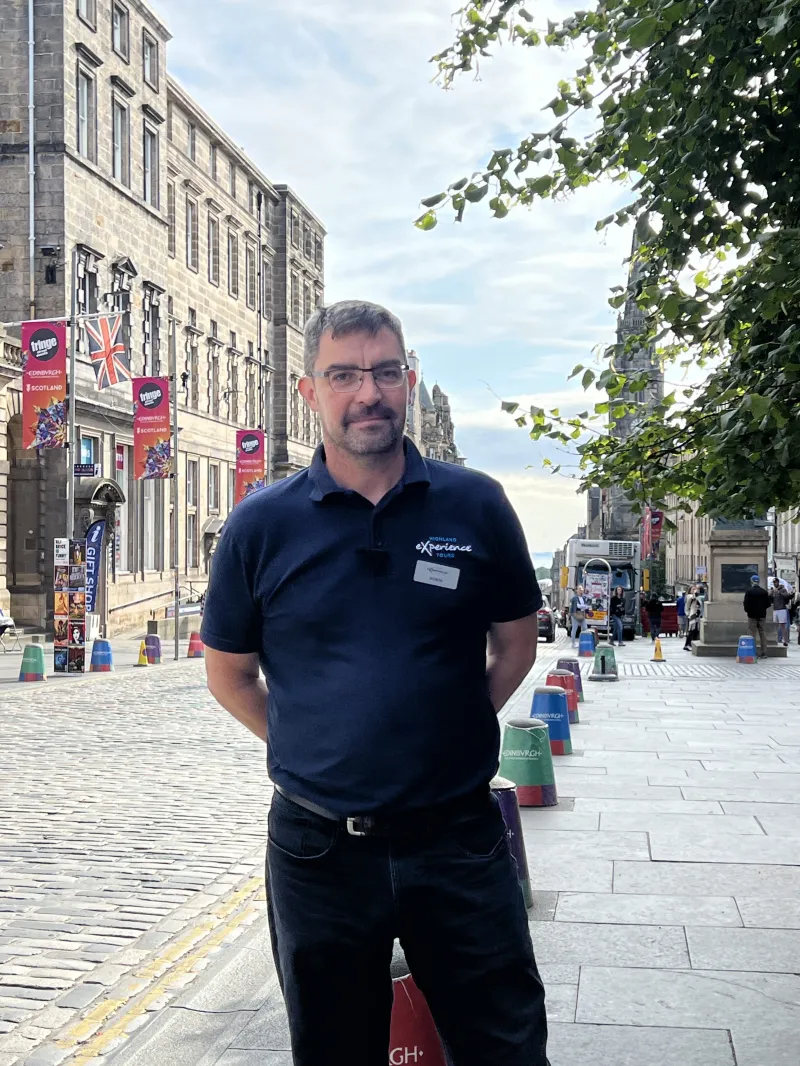 Man in a navy shirt stands on a cobblestone street lined with buildings and colorful banners.
