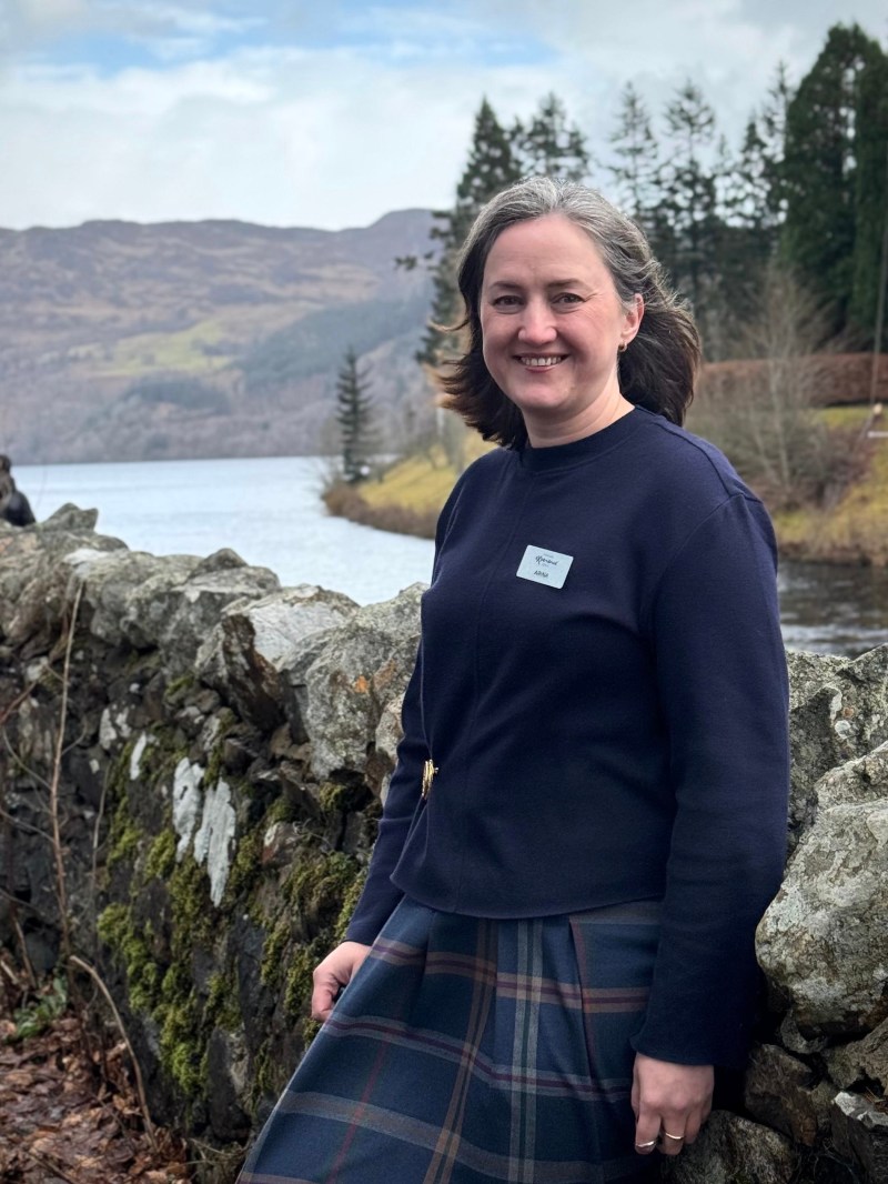Smiling woman in blue outfit standing by stone wall with scenic lake and hills in background.