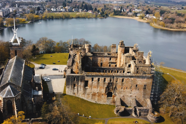 Aerial view of a historic stone castle by a lake with a church and a cemetery in the foreground.