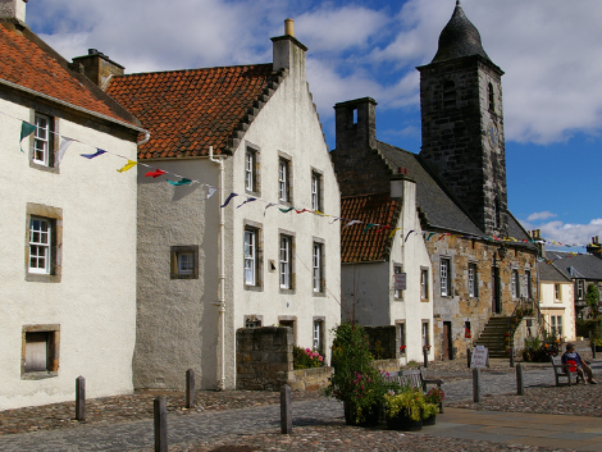 Historic village square with cobblestone paths, old buildings, and a church tower on a sunny day.