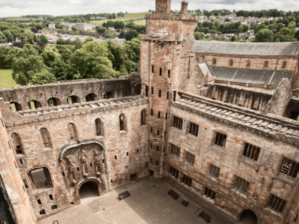 Historic stone castle courtyard with walls, tower, and arched entrances, surrounded by greenery and distant town.
