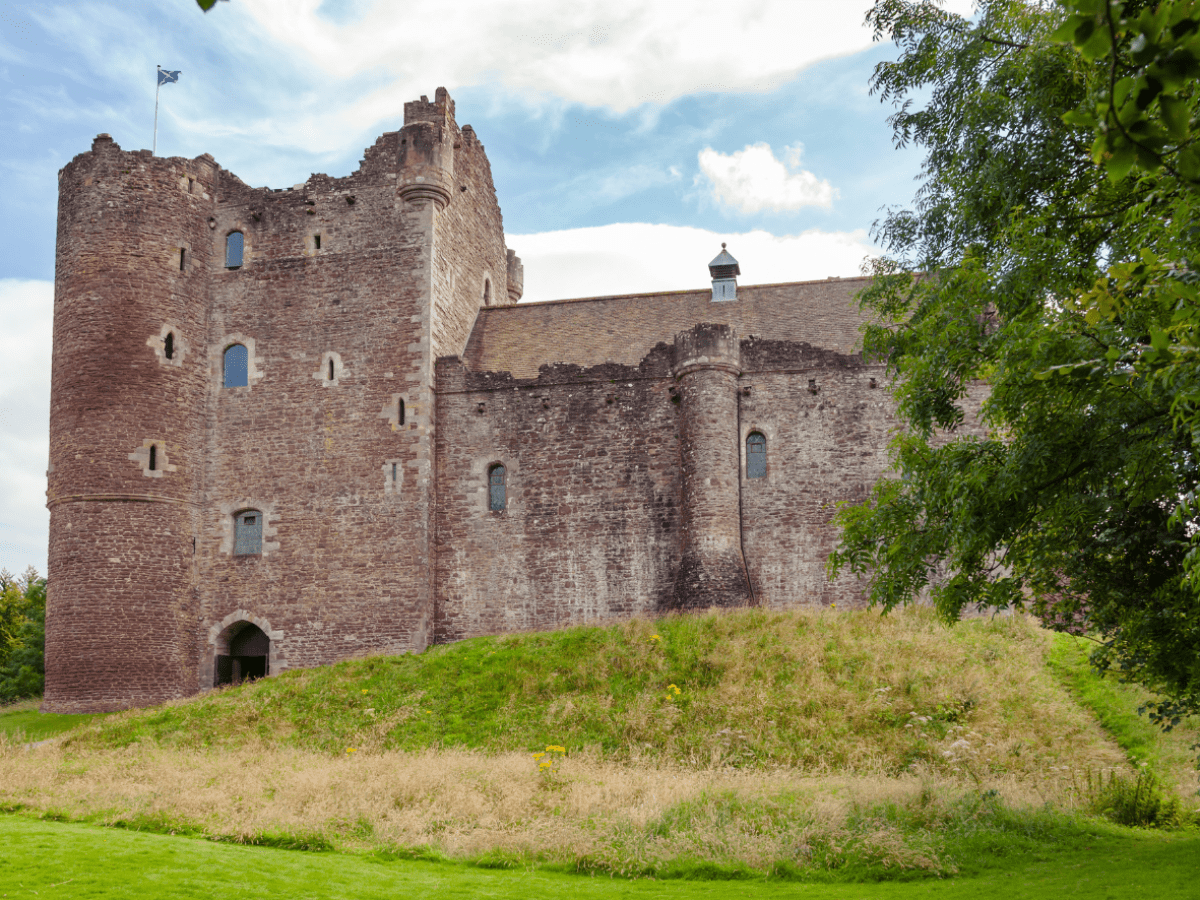 Medieval stone castle with turrets, surrounded by grass and trees under a cloudy sky.