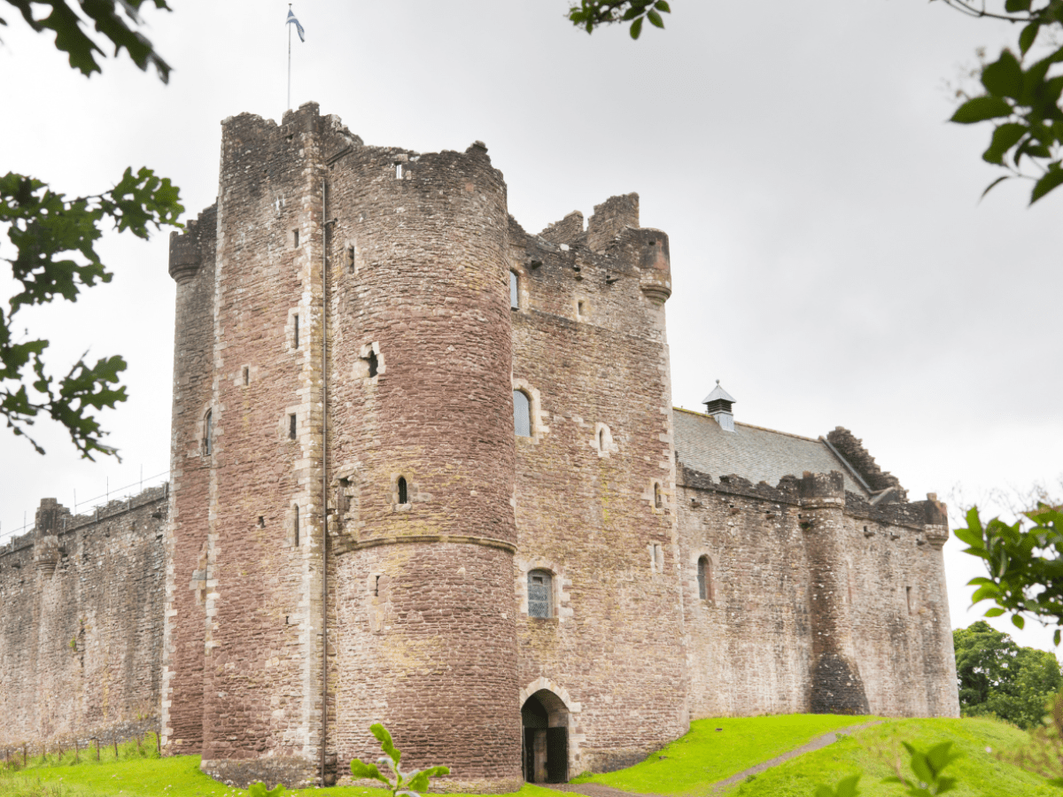 Medieval castle with a tower, surrounded by greenery and cloudy sky.