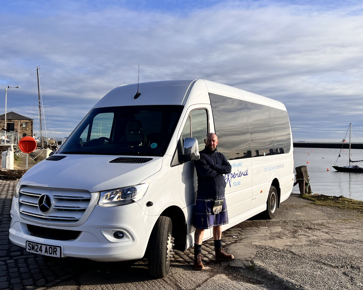 Man standing beside a white van near a harbor with boats.