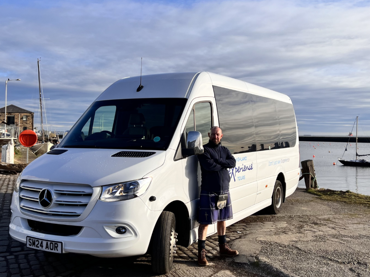 Man standing beside a white van near a harbor with boats.