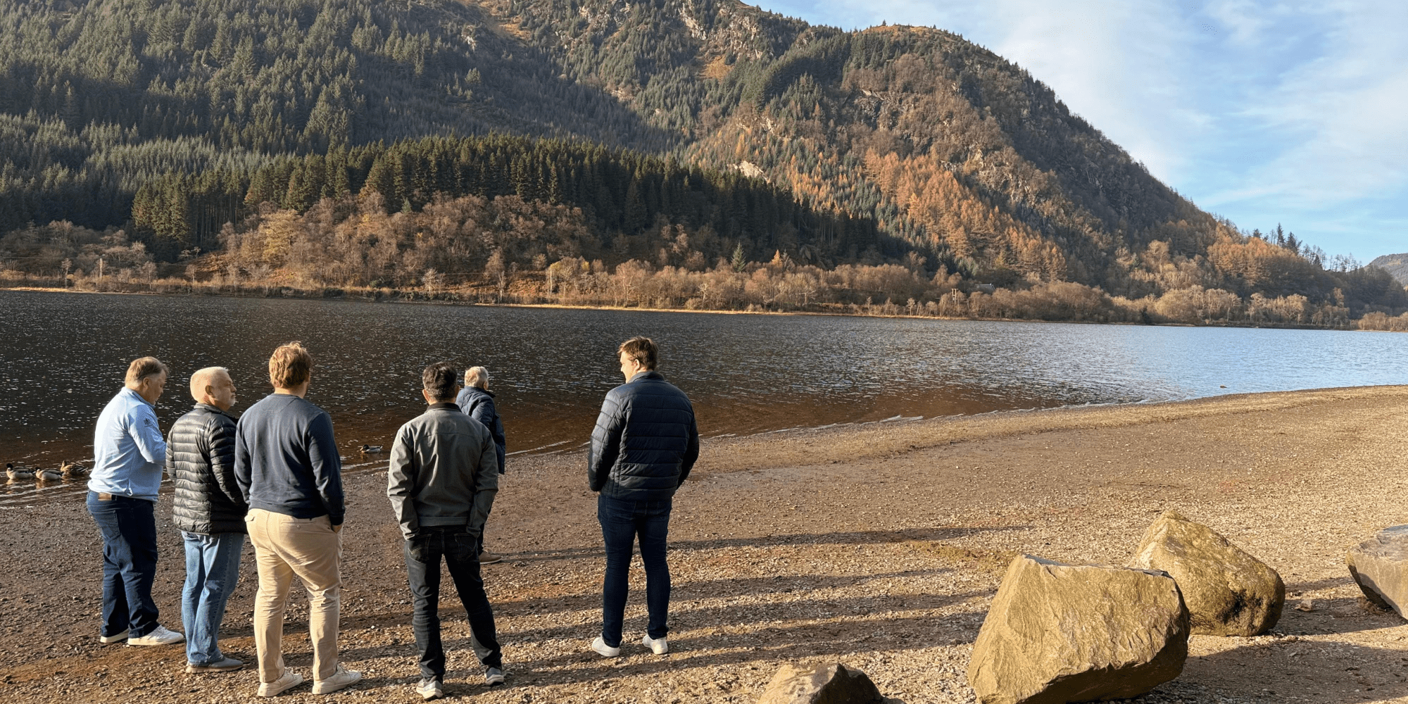 Group of people standing by a lake with wooded hills in the background.