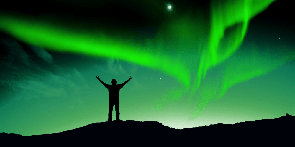 Silhouette of person with arms raised under bright green aurora lights in night sky.