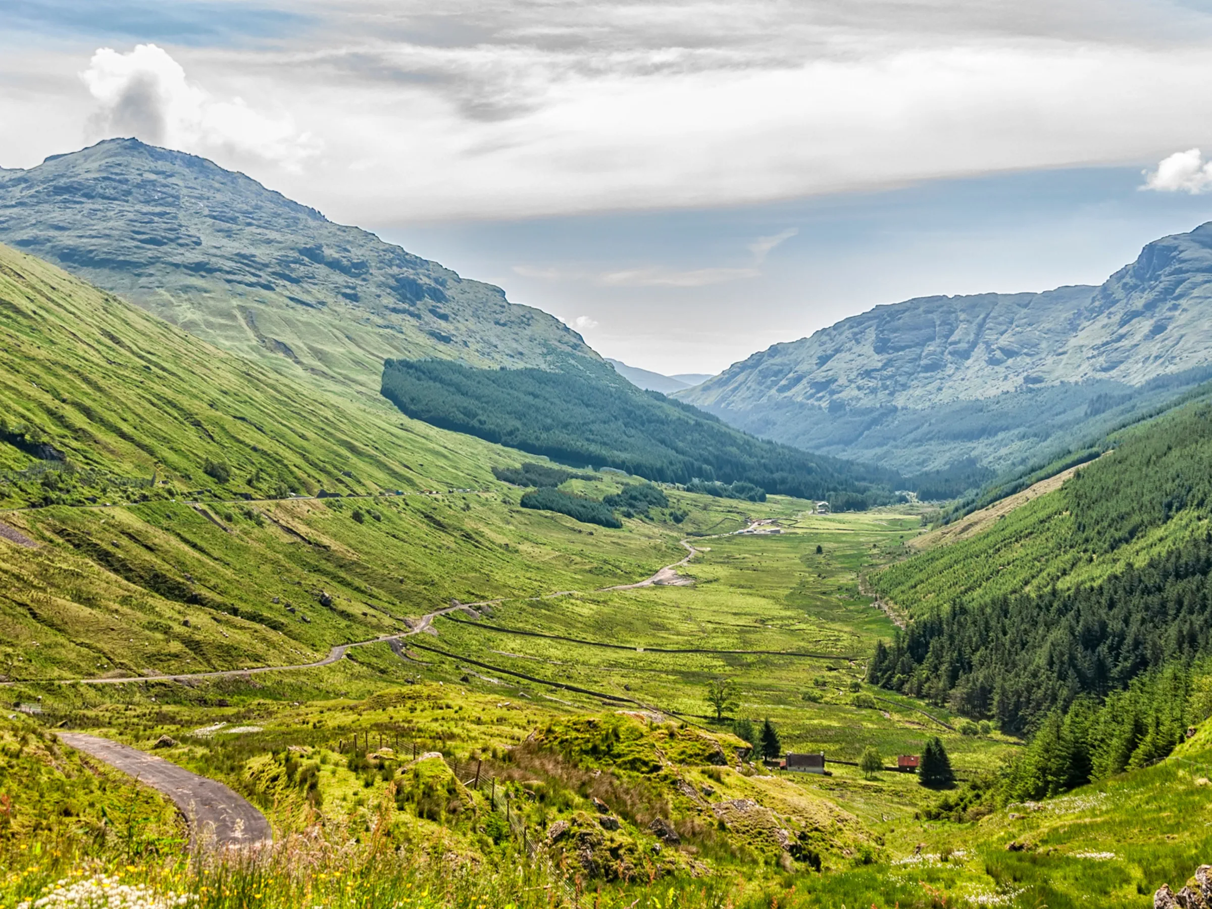 A lush valley with mountains, green slopes, and a cloudy sky in the background.