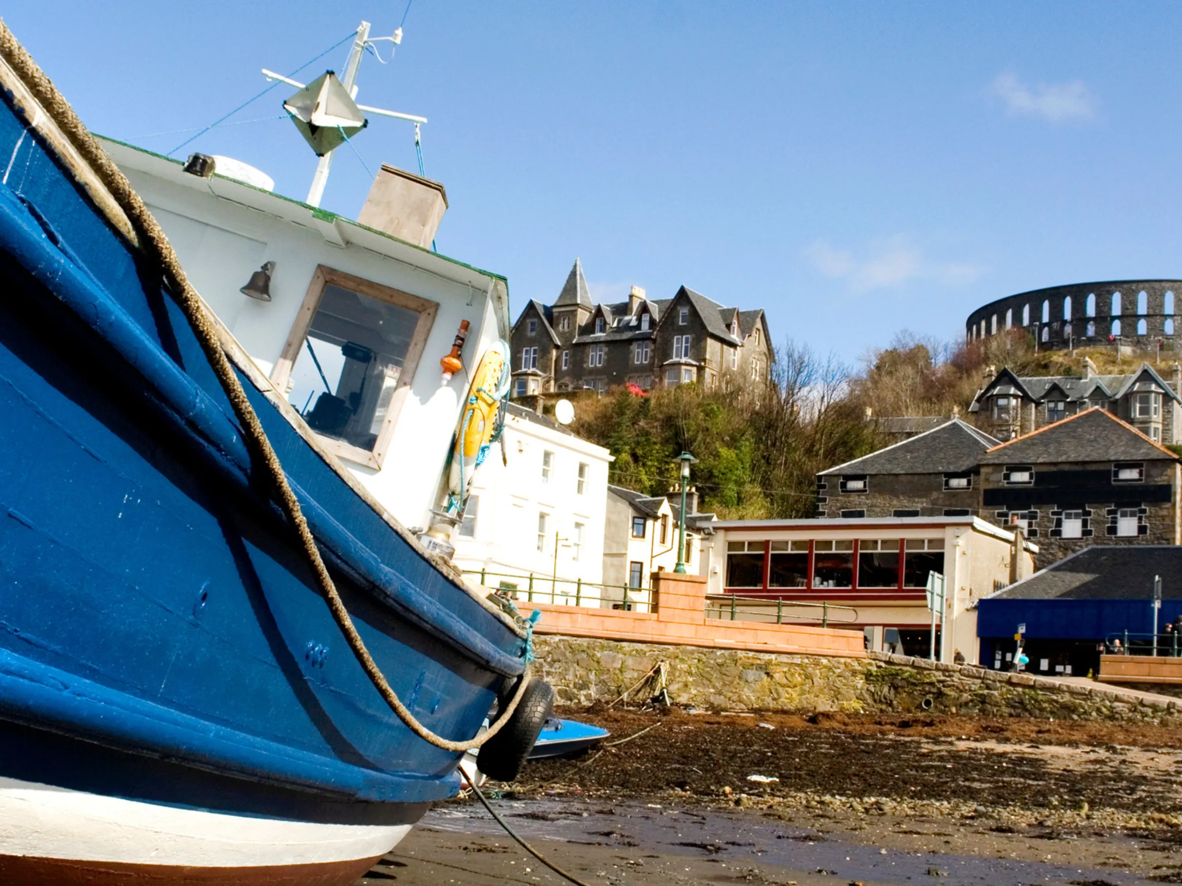 Blue boat on shore with town and large circular structure on hill in the background.