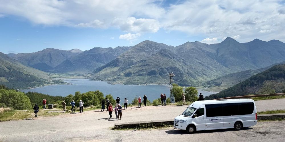 Tourists and a van at a viewpoint overlooking a lake and mountains.