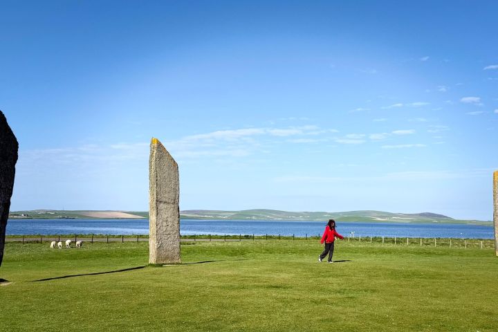 Person in red near three standing stones on a grassy field, with sheep and a lake in the background.