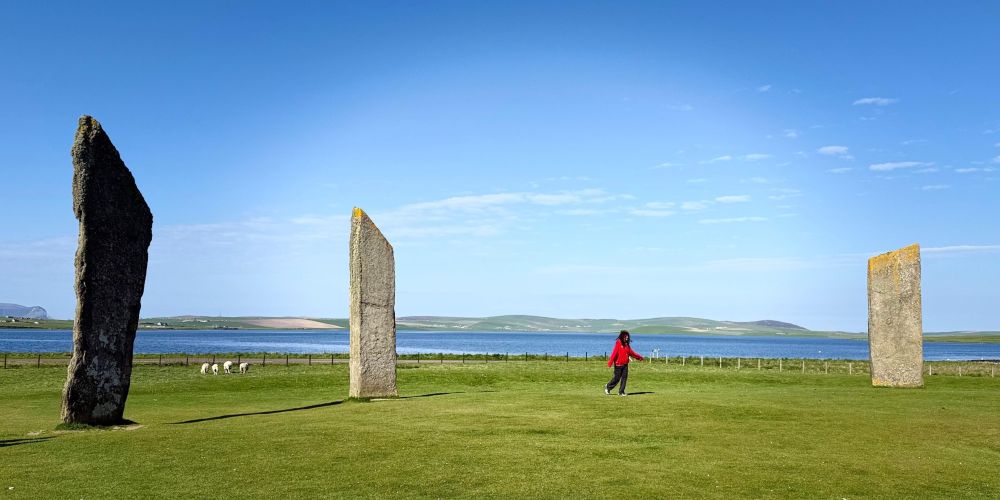 Person in red near three standing stones on a grassy field, with sheep and a lake in the background.