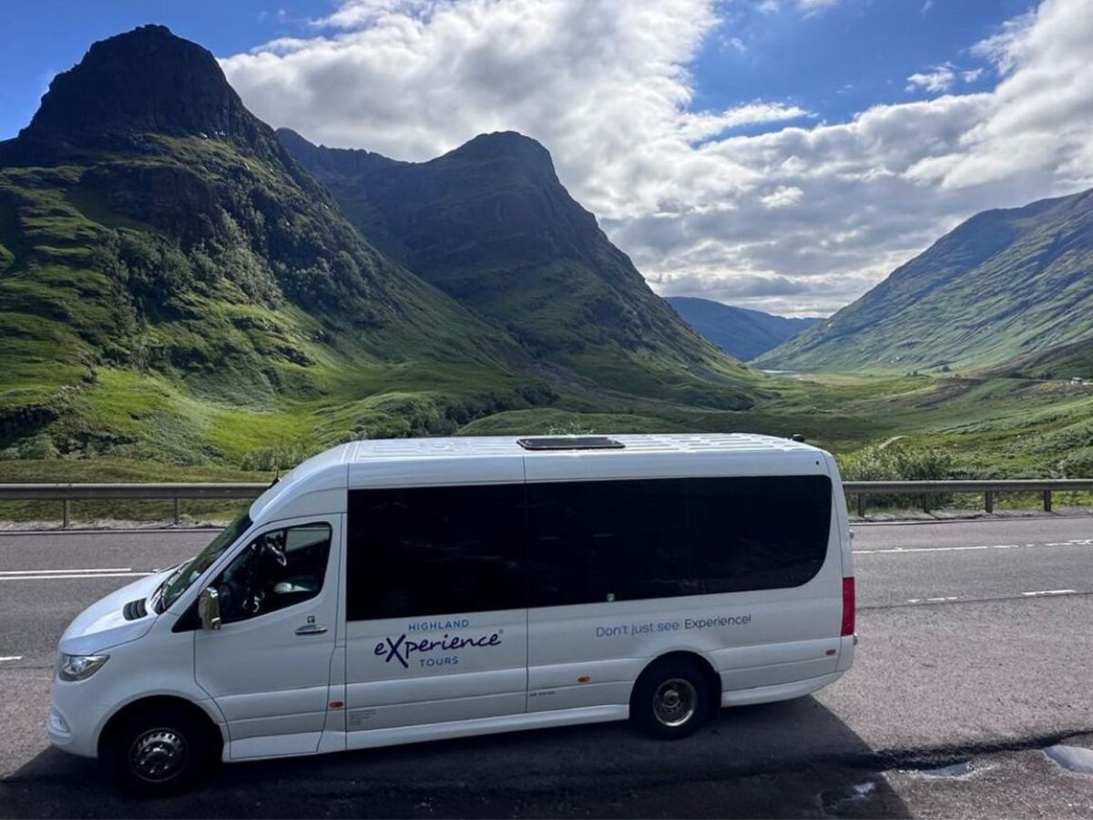 White tour bus on a road with mountainous landscape and cloudy sky in the background.