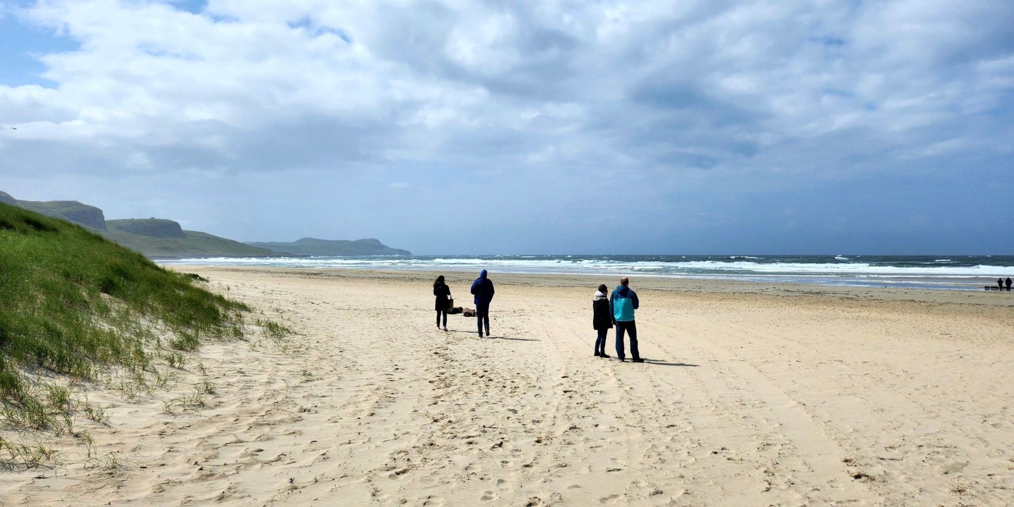 Four people walking on a beach with grass dunes on the left and cloudy sky.