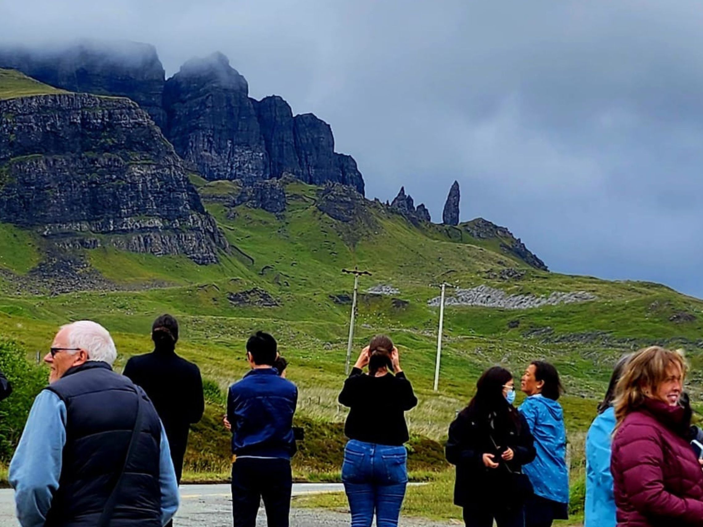 Group of people standing in front of a grassy mountain landscape with cloudy sky.