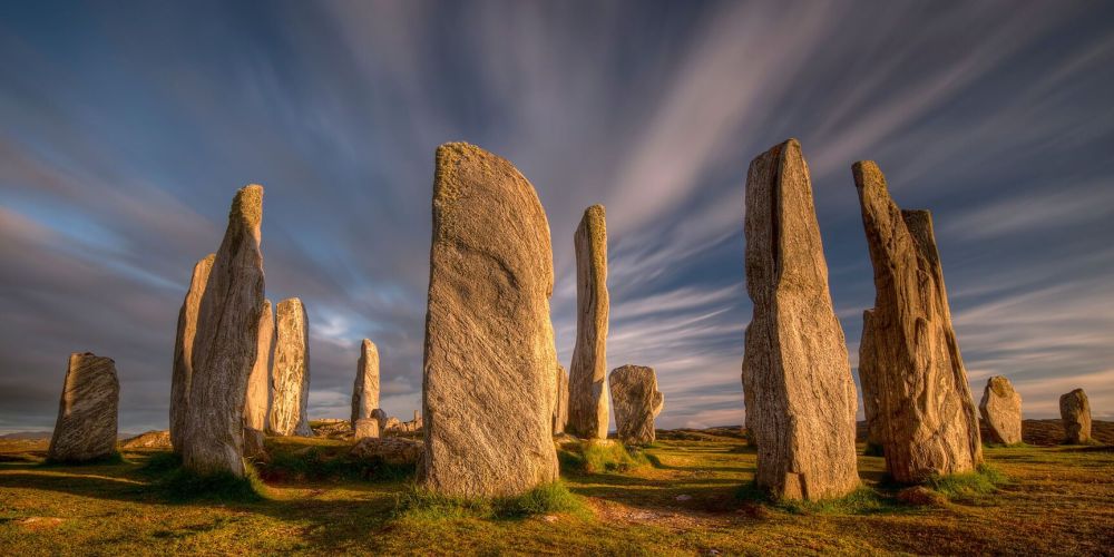 Tall standing stones under a cloudy sky, forming a circle on grassy terrain.