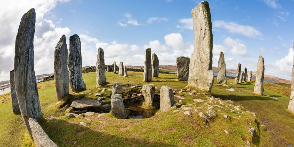Stone circle with tall standing stones on grassy terrain under a blue sky with clouds.