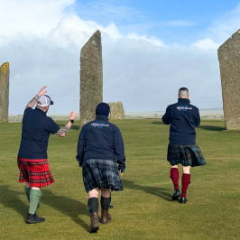 Three people in kilts walk towards standing stones on a grassy field under a mostly cloudy sky.