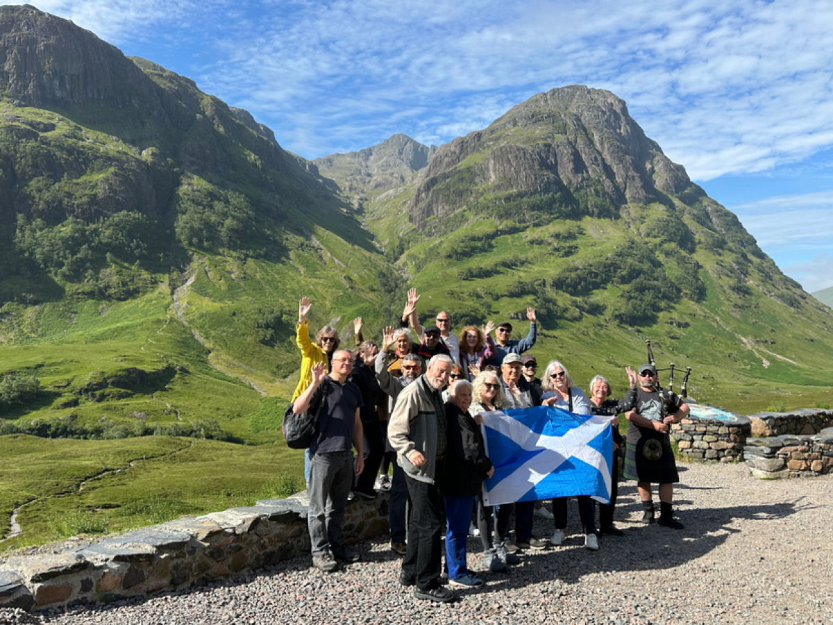 Group of people with Scottish flag and bagpiper in front of lush green mountains.