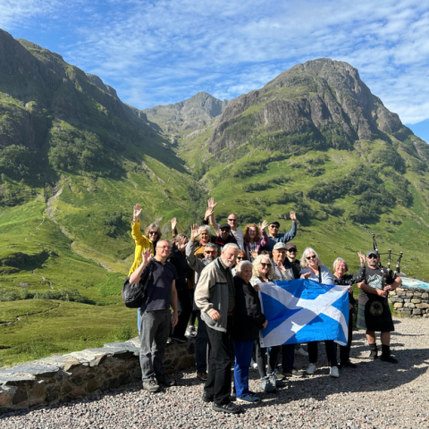 Group of people with Scottish flag and bagpiper in front of lush green mountains.