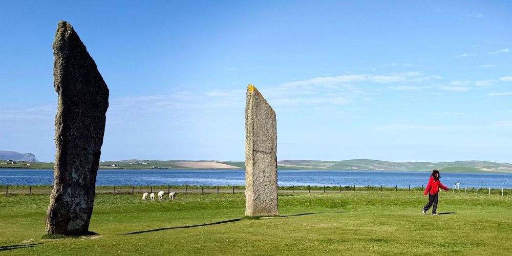 Person in red near standing stones, with sheep and a lake in the background under a blue sky.