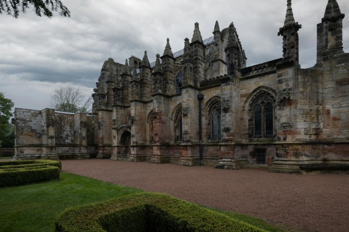 Gothic stone chapel with ornate architecture, cloudy sky, green hedges in foreground.