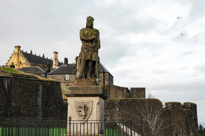 Statue of a historical figure with castle walls and building in the background, under a cloudy sky.