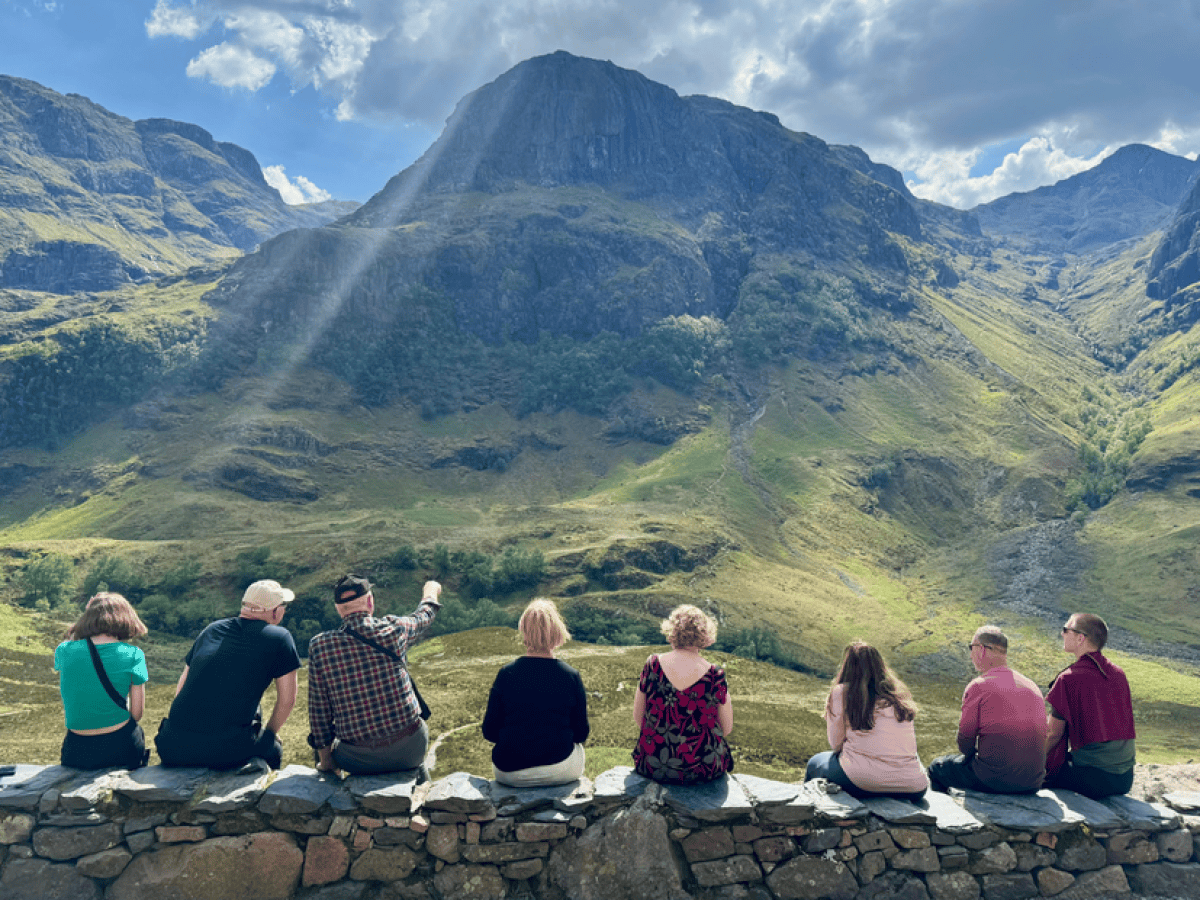 Group of seven people sitting on a stone wall, viewing a mountainous landscape with sunlight streaming down.