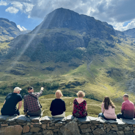 Group of seven people sitting on a stone wall, viewing a mountainous landscape with sunlight streaming down.