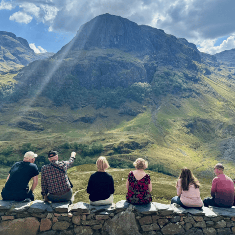 Group of seven people sitting on a stone wall, viewing a mountainous landscape with sunlight streaming down.
