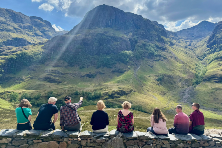Group of seven people sitting on a stone wall, viewing a mountainous landscape with sunlight streaming down.