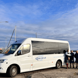 White tour van parked by water, group of people standing beside it on a sunny day.