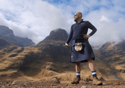 Man stands in front of rugged mountains wearing a blue kilt and hiking boots.