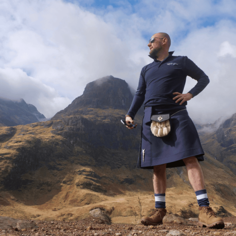 Man stands in front of rugged mountains wearing a blue kilt and hiking boots.