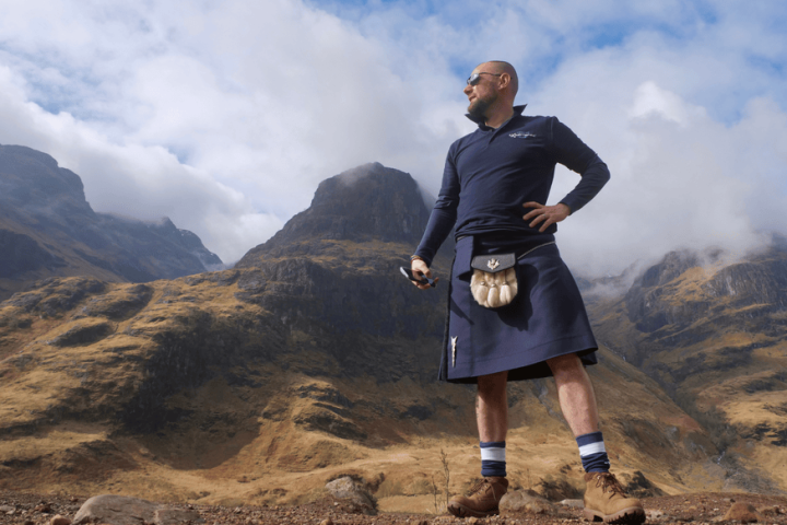 Man stands in front of rugged mountains wearing a blue kilt and hiking boots.