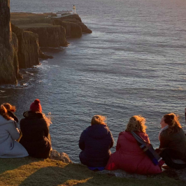 Group of people sitting by a cliff at sunset with a lighthouse in the background.