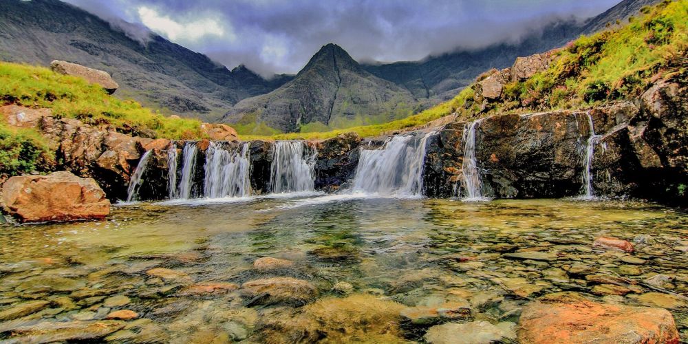 Small waterfall with clear water and rocky landscape, mountains in the background, under a cloudy sky.