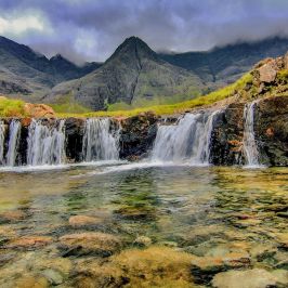 Small waterfall with clear water and rocky landscape, mountains in the background, under a cloudy sky.