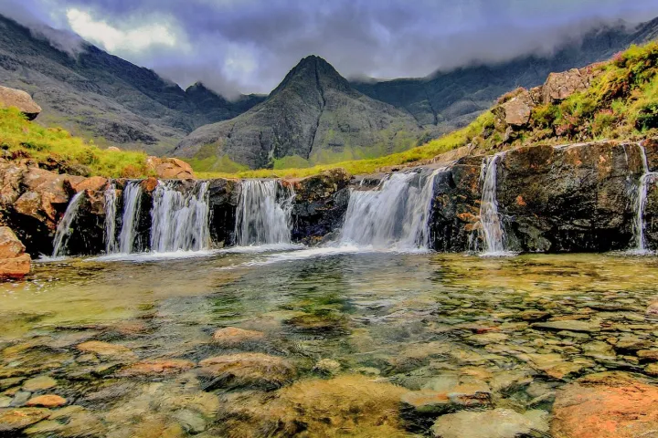 Small waterfall with clear water and rocky landscape, mountains in the background, under a cloudy sky.