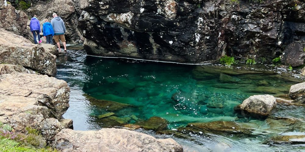 People walking near a clear blue rock pool surrounded by rugged rocks.