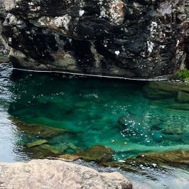 People walking near a clear blue rock pool surrounded by rugged rocks.