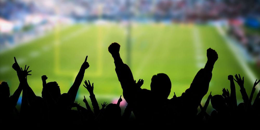 Silhouettes of fans cheering at a football stadium with a blurred field in the background.