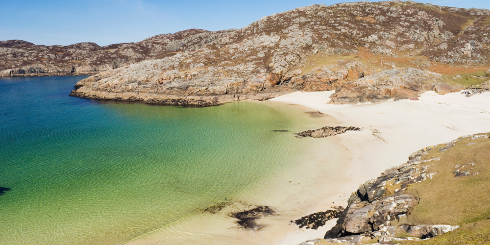 Sandy beach with clear water and rocky hills under a blue sky.