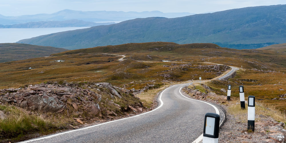 Winding road through hilly landscape with distant mountains and cloudy sky.