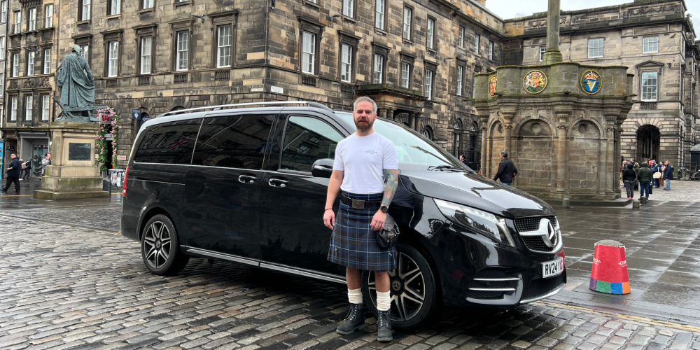 Man in kilt stands by black van on cobblestone street, historic buildings in background.