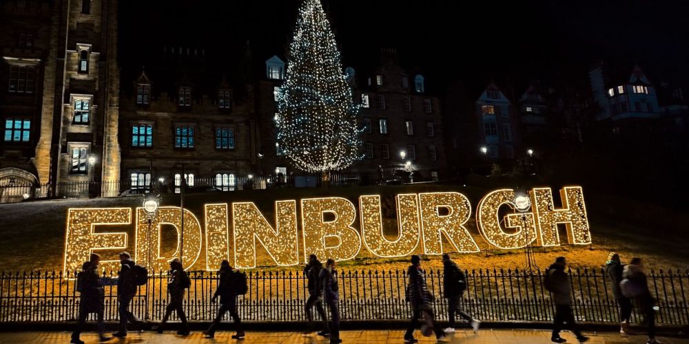 People walk past illuminated 'EDINBURGH' sign and Christmas tree at night.