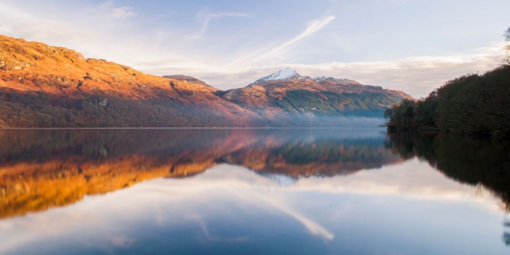 Calm lake reflecting autumn hills and a snow-capped mountain under a clear sky.