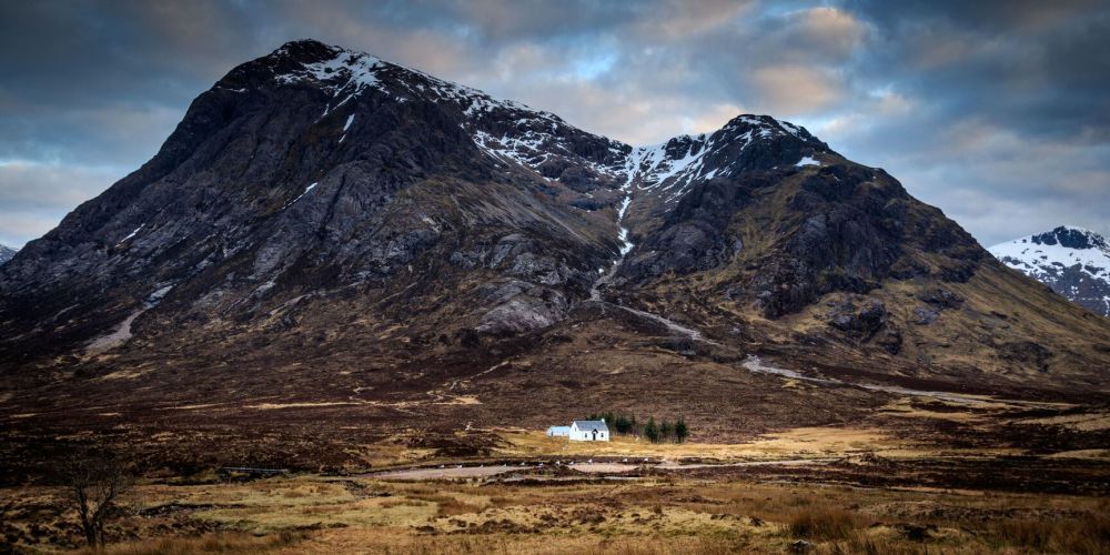 White cottage under towering mountain with snow patches and cloudy sky.
