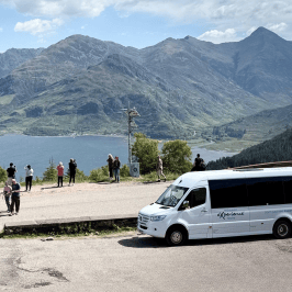 Group of people near a parked van, overlooking mountains and a large lake.