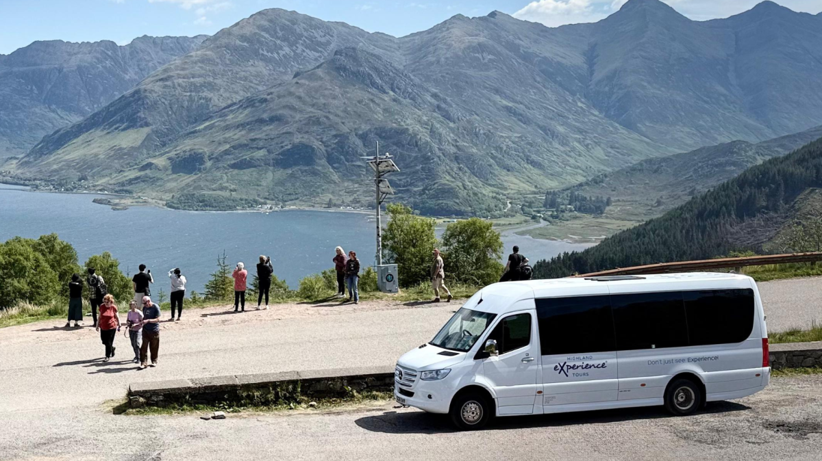 Group of people near a parked van, overlooking mountains and a large lake.